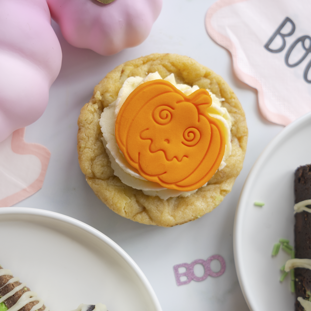 A Halloween-themed cookie featuring a golden-brown cookie base, white frosting, and a bright orange pumpkin-shaped decoration with a carved face. The cupcake is styled on a white surface with pink decorative props and partial plates of food.