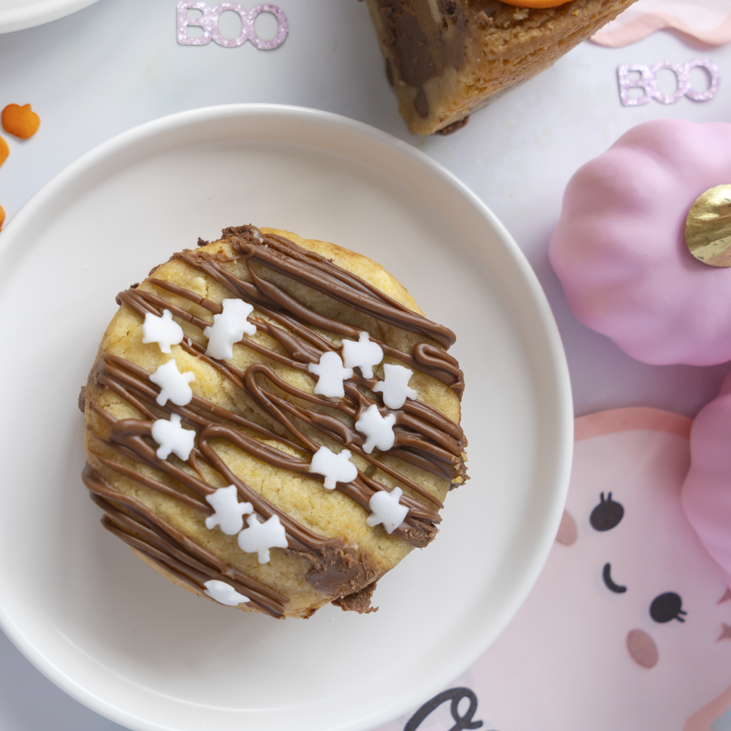 A round pumpkin spice cookie topped with chocolate drizzle and white icing stars, presented on a white plate with Halloween-themed props in the background.