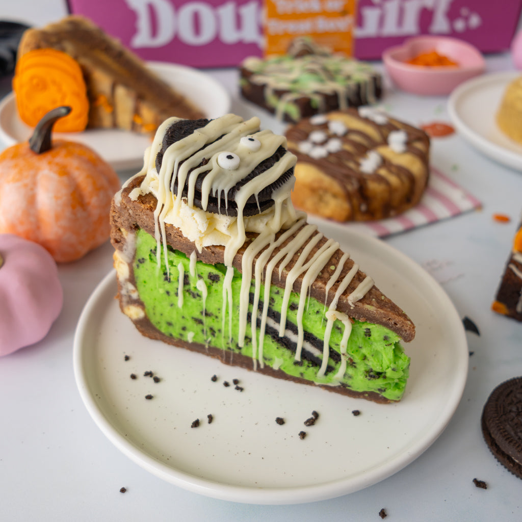 A Halloween-themed slice of cookie pie on a white plate, decorated with green frosting, white drizzle, and an Oreo cookie on top, surrounded by pumpkins and other decorated cakes.