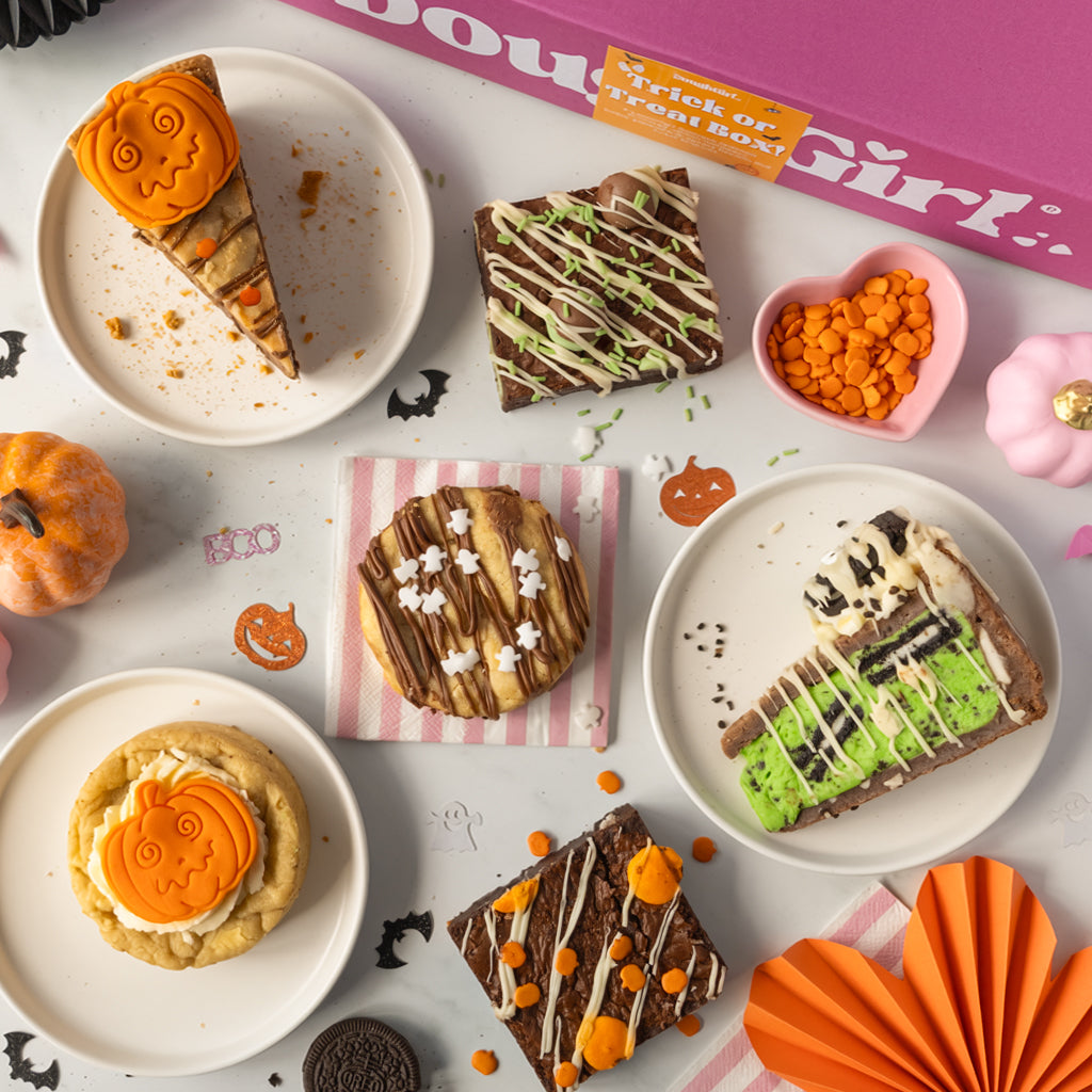 A styled flat-lay of Halloween-themed dessert treats on white plates, including pumpkin cake slices, chocolate brownies with drizzles, and cookies, arranged with orange paper fans, candy corn, a pink heart dish, and a pink box labeled 'DoughGirl' on a white surface.