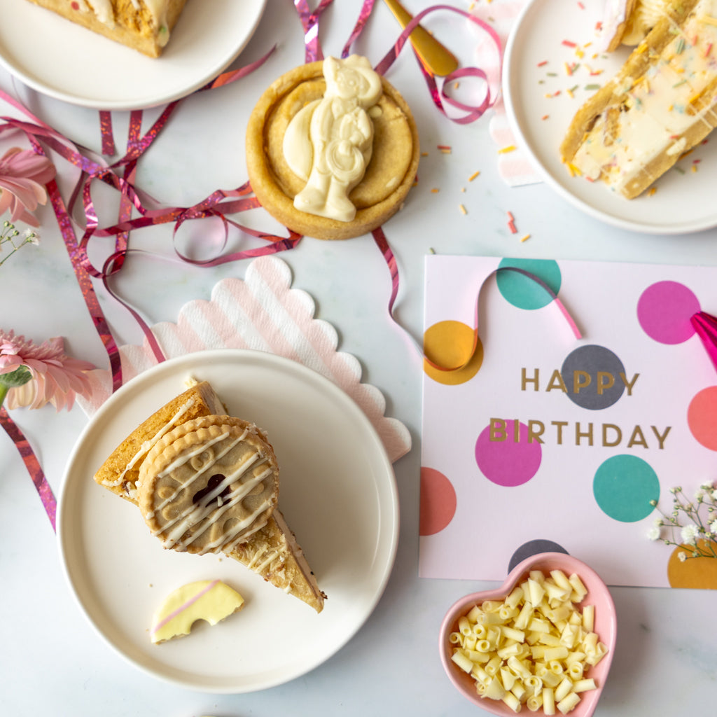 A styled flat-lay of assorted baked goods and party decorations on a light marble surface. Slices of cookie pie and cookies are arranged on white plates, with pink ribbons, a small bowl of macaroni candy, and a pink and gold 'HAPPY BIRTHDAY' card on a white background.