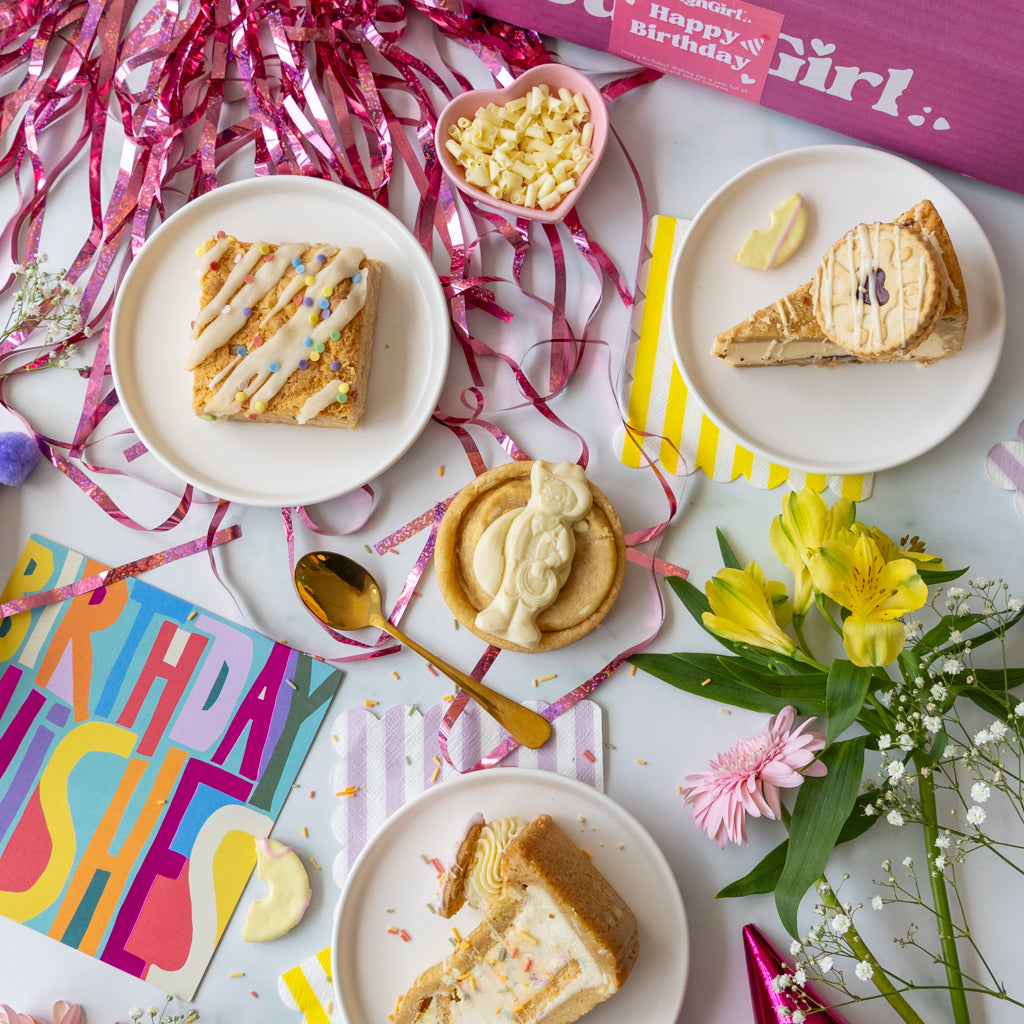 A styled birthday dessert spread with four plates of cake slices, a small bowl of white candies, a gold spoon, colorful birthday chocolate curls, pink tinsel, flowers, and a pink 'Happy Birthday DoughGirl' box on a white surface.