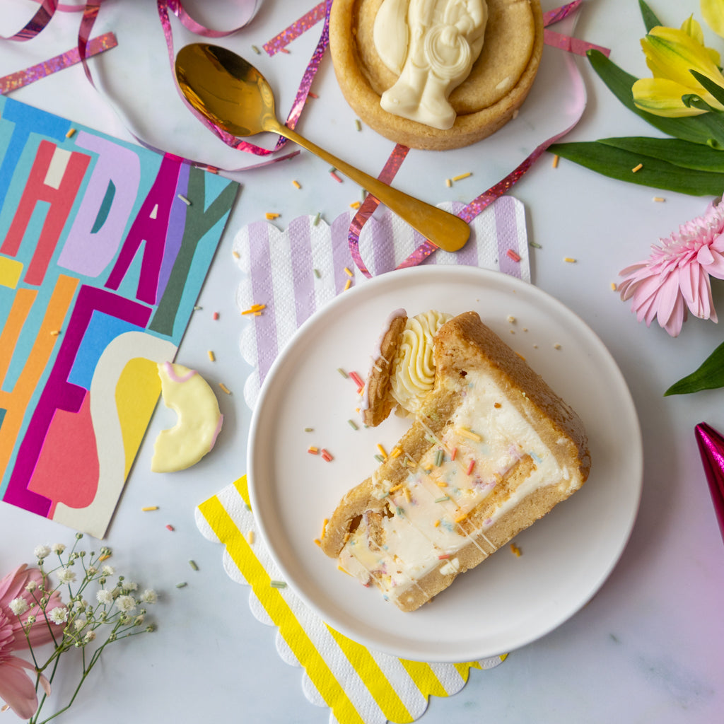 A slice of cookie pie on a white plate, styled in a colorful flat-lay with a gold spoon, a birthday card, flowers, and pastel napkins on a light marble surface.