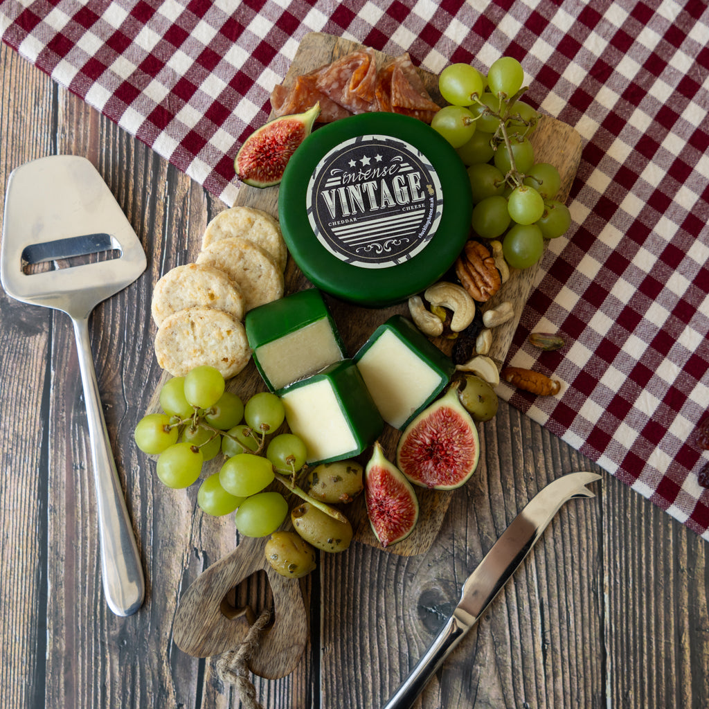 A styled charcuterie board featuring a round green cheese labeled 'INTENSE VINTAGE', alongside sliced cheeses, crackers, green grapes, figs, nuts, and a cheese knife on a rustic wooden table with a red-and-white checkered cloth.