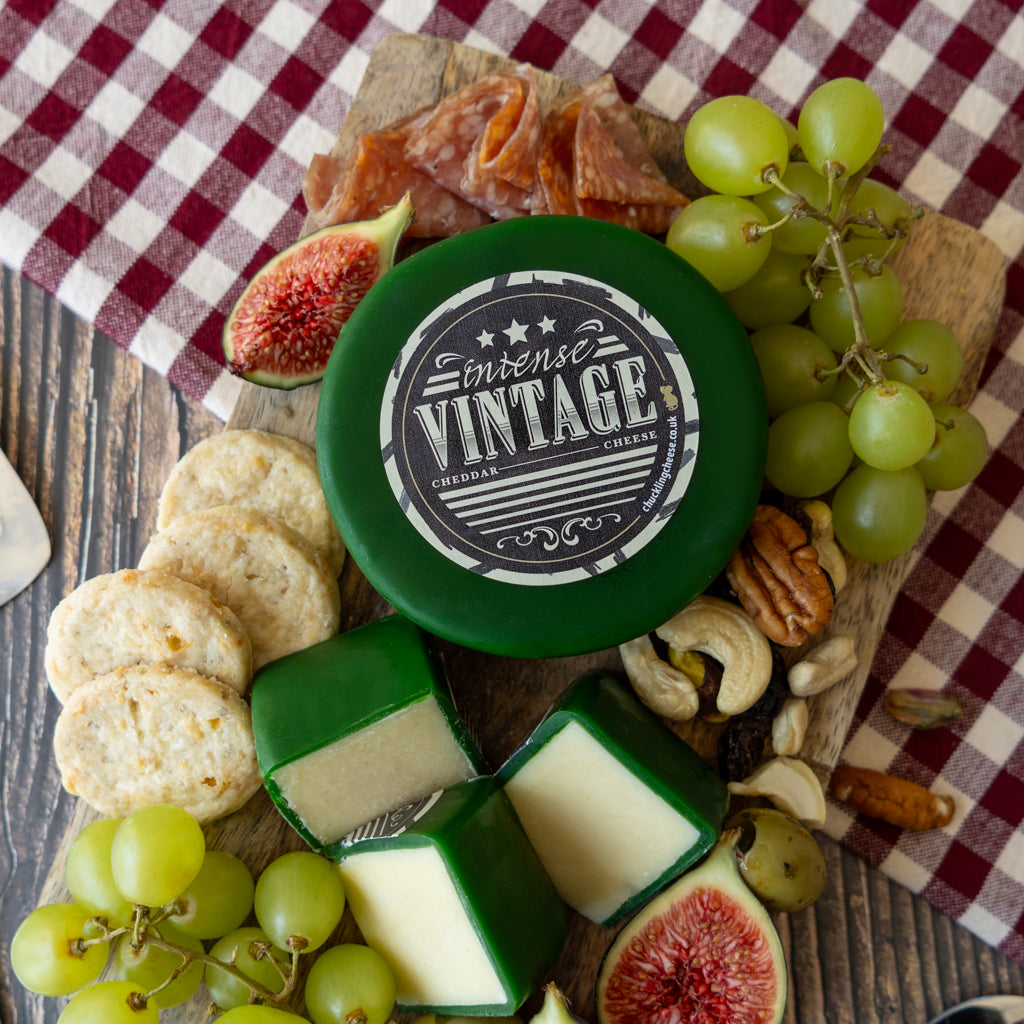 A round wheel of Intense Vintage Cheese with a dark green rind, styled on a wooden board with crackers, figs, green grapes, walnuts, pecans, and sliced prosciutto on a red and white checkered cloth.