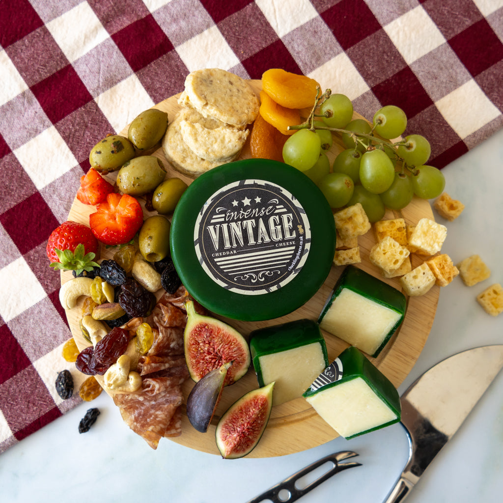 A styled charcuterie board with an assortment of cheeses, olives, grapes, figs, crackers, nuts, and dried fruit, centered around a round green cheese container labeled 'Vintage Cheddar Cheese'.