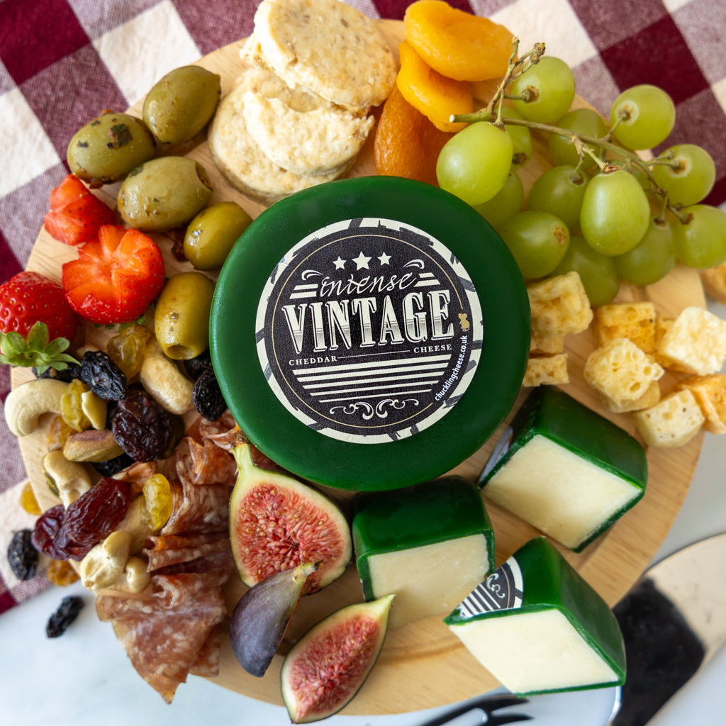 A round wooden charcuterie board filled with cheese, olives, grapes, figs, strawberries, crackers, nuts, and prosciutto, centered around a round green cheese wheel labeled 'intense VINTAGE CHEDDAR CHEESE'.