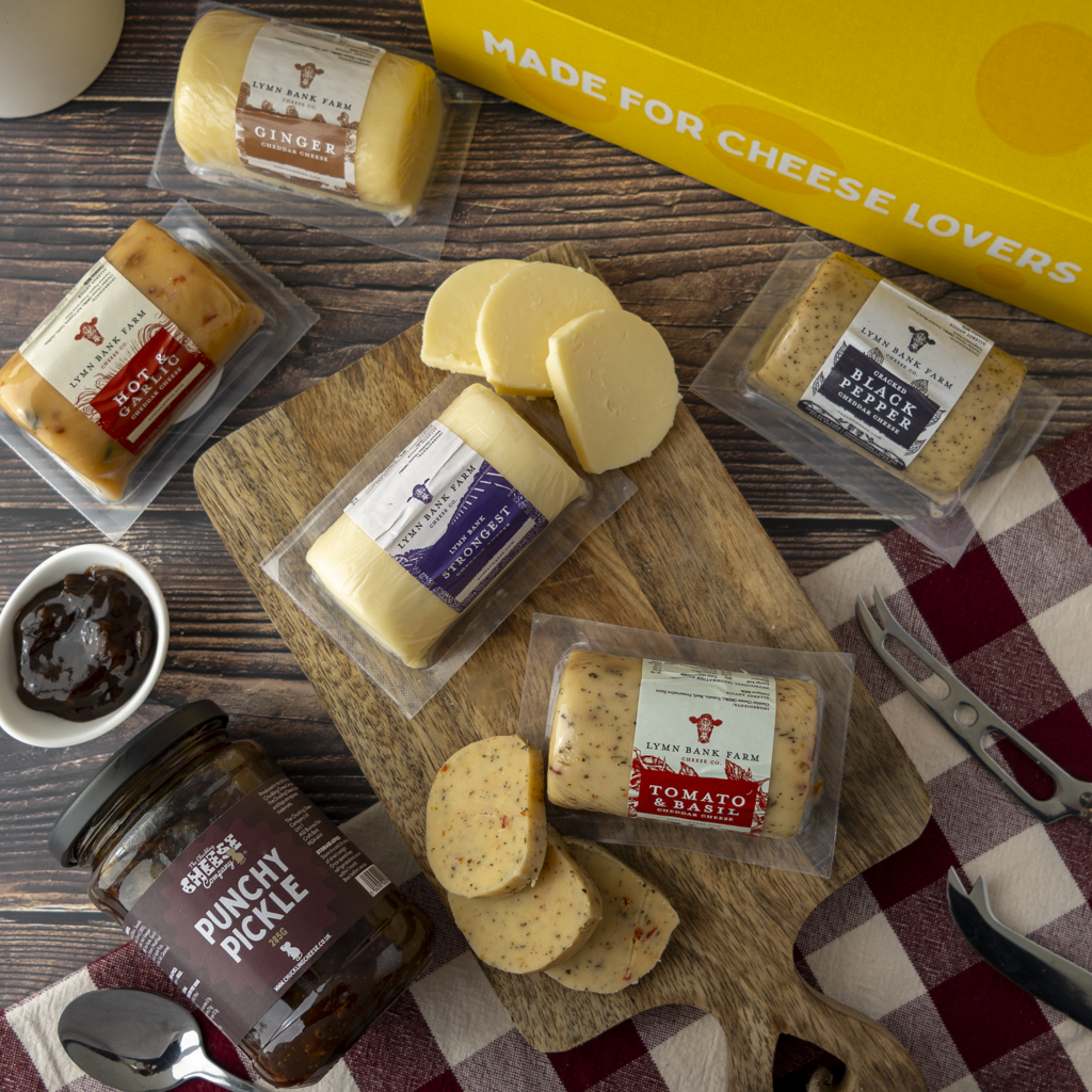 A styled cheese and charcuterie spread on a wooden board with a yellow 'Made for Cheese Lovers' box, multiple packaged cheeses, sliced cheeses, a jar of punchy pickle, scissors, and a red-and-white check cloth on a wood surface.