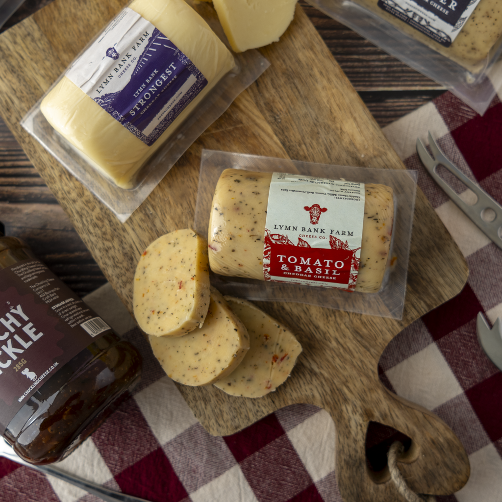 Lynn Bank Farm tomato & basil cheese barrels and a packaged block on a wooden cutting board with sliced cheese, a bottle of chilli oil, and a red-and-white checkered cloth in the background.