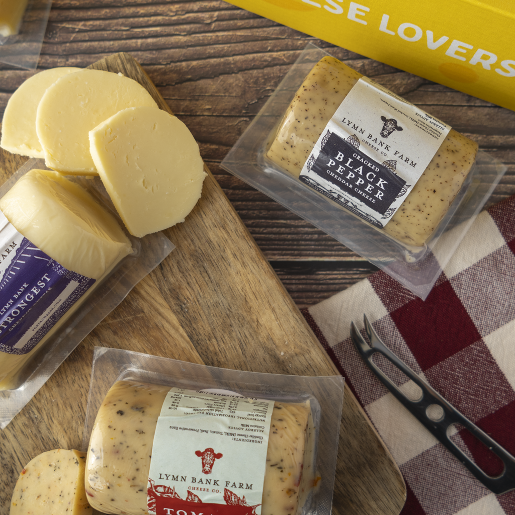 Three packages of Lymn Bank Farm artisanal cheeses arranged on a wooden cutting board with sliced cheese pieces, alongside a black cheese knife and a red-and-white checkered cloth on a rustic wooden table.