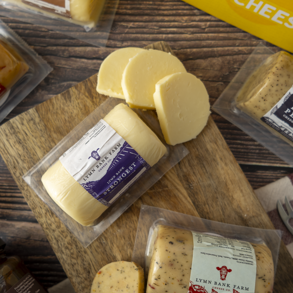 Several packages of Lymn Bank Farm cheese barrels and slices are arranged on a wooden cutting board over a dark wood tabletop. The cheese is pale yellow with some visible speckling, and the labels show Lymn Bank Farm branding.