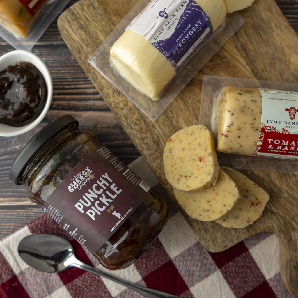 A styled food product flat lay featuring Lymn Bank Cheese products, including a jar of Punchy Pickle, packaged cheese wedges, and sliced cheese on a wooden cutting board with a spoon and checkered cloth.