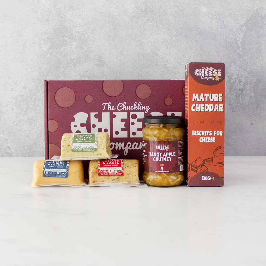 A cheese gift set from The Chuckling Cheese Company, featuring a red gift box, a jar of chutney, a box of mature cheddar, and three wrapped cheese barrels, arranged on a white surface against a textured grey background.