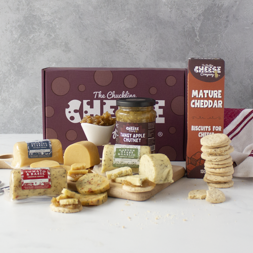 A cheese and biscuit gift assortment displayed on a white surface against a grey wall. The set includes a dark burgundy box labelled 'The Chuckling Cheese', a jar of Randy Apple Chutney, a box of Mature Cheddar from Cheese Company, and an assortment of cheeses, crackers, and biscuits arranged in front.
