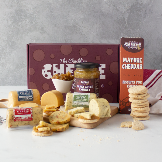 A cheese and biscuit gift assortment displayed on a white surface against a grey wall. The set includes a dark burgundy box labelled 'The Chuckling Cheese', a jar of Randy Apple Chutney, a box of Mature Cheddar from Cheese Company, and an assortment of cheeses, crackers, and biscuits arranged in front.