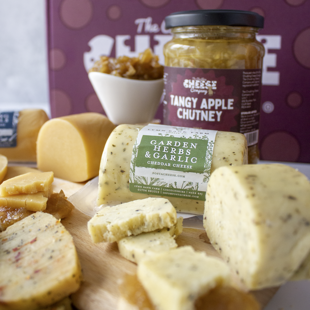 A styled food product display featuring a wheel and slices of cheddar cheese with a 'Garden Herbs & Garlic' label, alongside a jar of 'Tangy Apple Chutney' and other cheese pieces on a wooden board, with a branded purple box in the background.