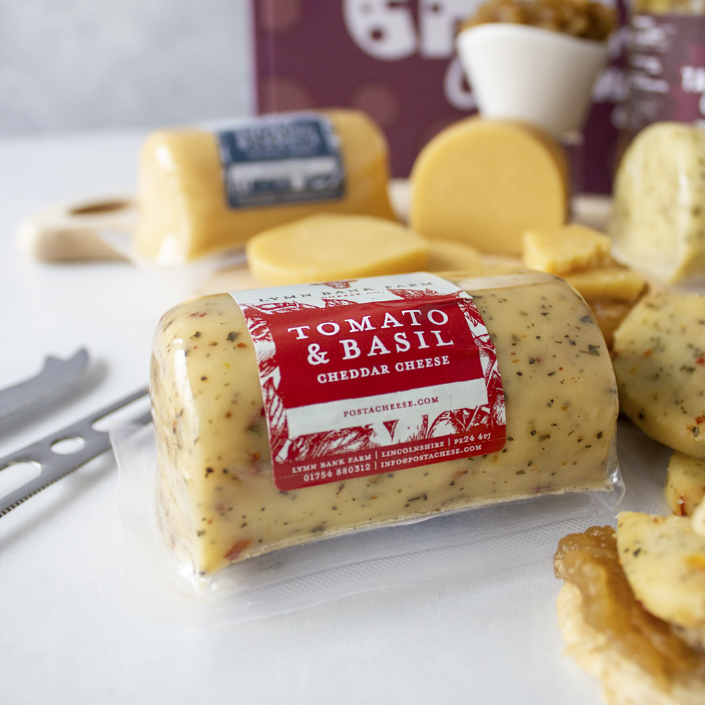 A barrel of Tomato & Basil Cheddar Cheese is shown in the foreground on a white surface, with sliced cheese and crackers arranged around it in a styled food setting.