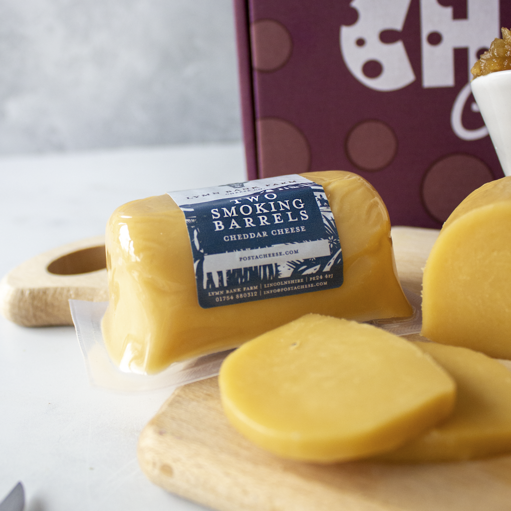 A package of Two Smoking Barrels Cheddar Cheese, shown with sliced cheese on wooden boards and a branded box in the background.