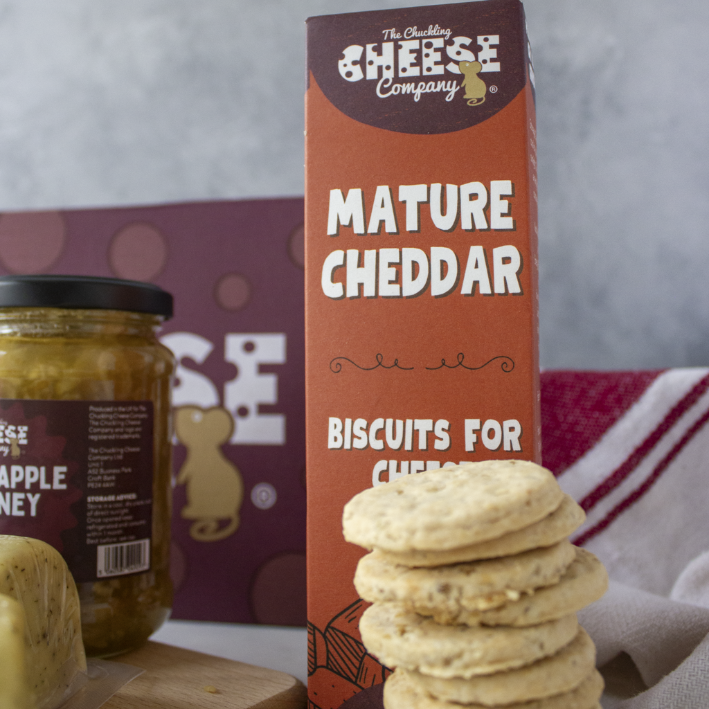 A box of The Chuckling Cheese Company Mature Cheddar Biscuits for Cheese, styled with a jar of cheddar apple honey and a stack of biscuits in the foreground.