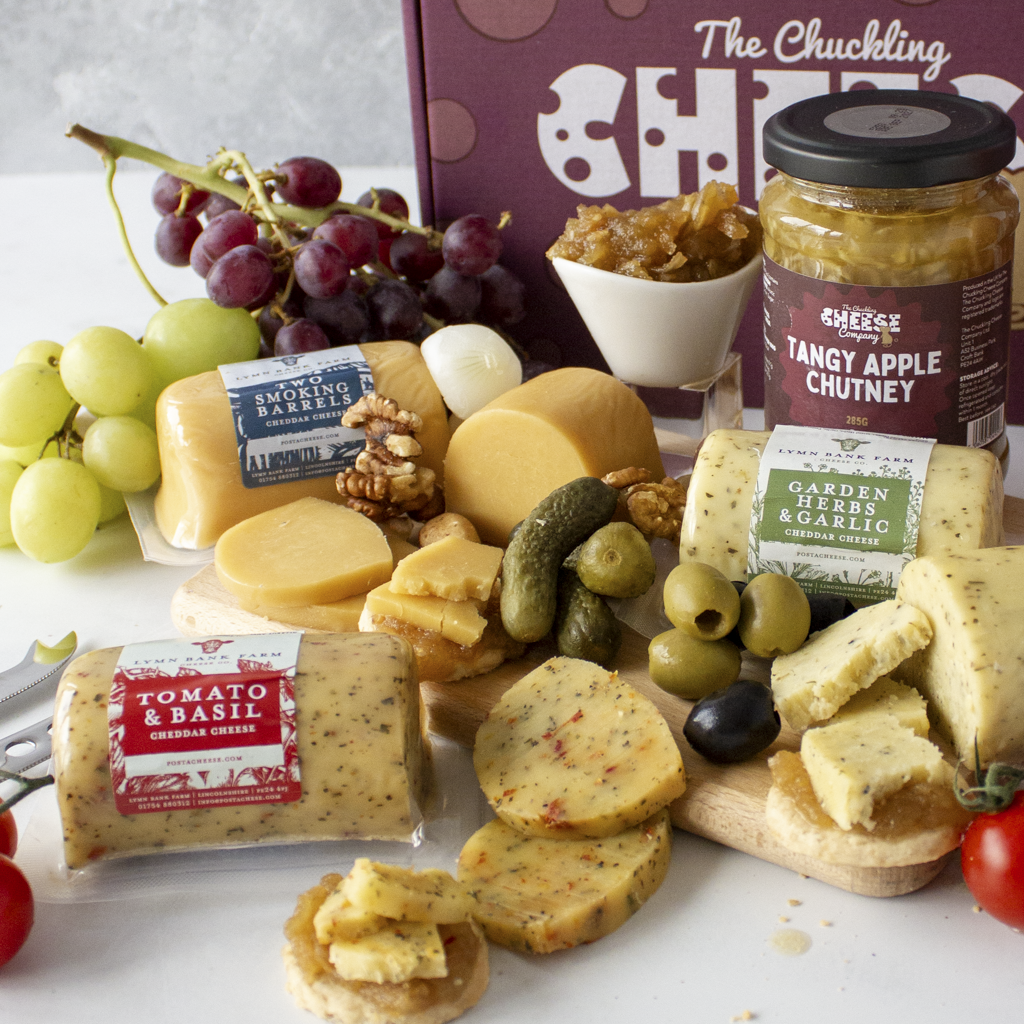 A gourmet cheese and charcuterie board arrangement on a white surface, featuring assorted cheeses, sliced tomatoes, grapes, olives, pickles, chutney, crackers, and a large purple gift box labeled 'The Chuckling Cheese Co.'.
