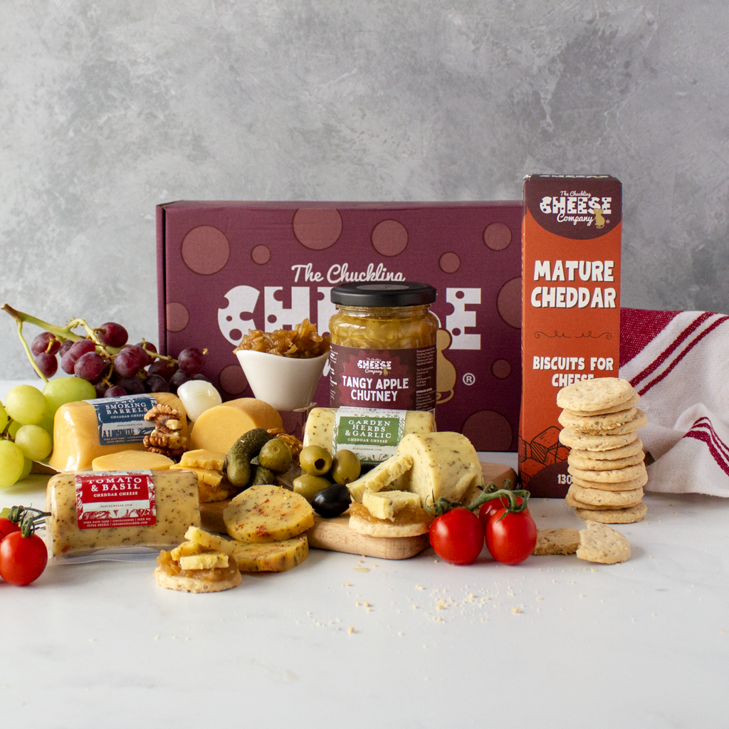 A gourmet cheese and charcuterie gift spread arranged on a white surface against a textured grey background. Visible products include a large burgundy gift box labelled 'The Chuckling Cheese', a jar of tangy apple chutney, a box of mature cheddar biscuits, and sliced cheeses, olives, grapes, and cherry tomatoes.