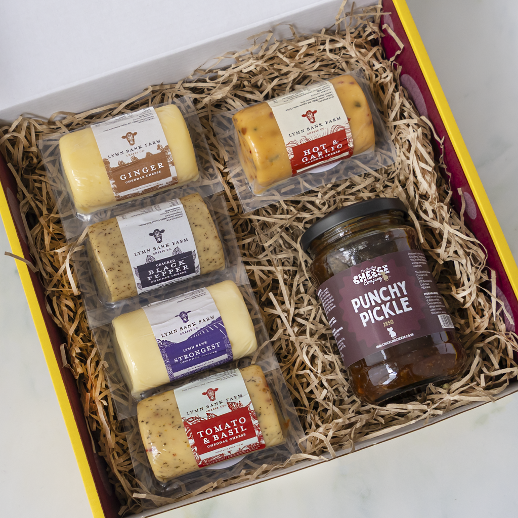 A gift box of cheese and pickle from The Chuckling Cheese Company, filled with shredded paper and containing six cheese barrels in clear plastic packaging and a jar of punchy pickle.