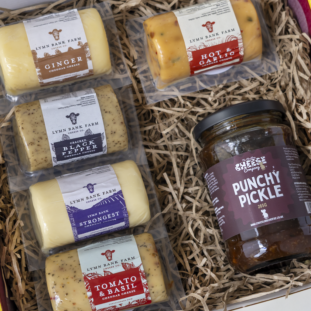 A gift box arrangement featuring six packages of Lymn Bank Farm artisanal cheeses and a glass jar of The Chuckling Cheese Company Punchy Pickle, set in a box filled with shredded paper.