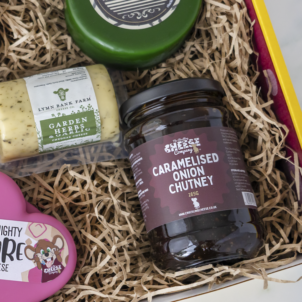 A curated cheese and chutney gift box filled with shredded paper. Visible items include a jar of Caramelised Onion Chutney from The Chuckling Cheese Company, a package of Lynn Bank Farm Garden Herb Cheese, a pink heart-shaped cheese tin, and a green round cheese piece.