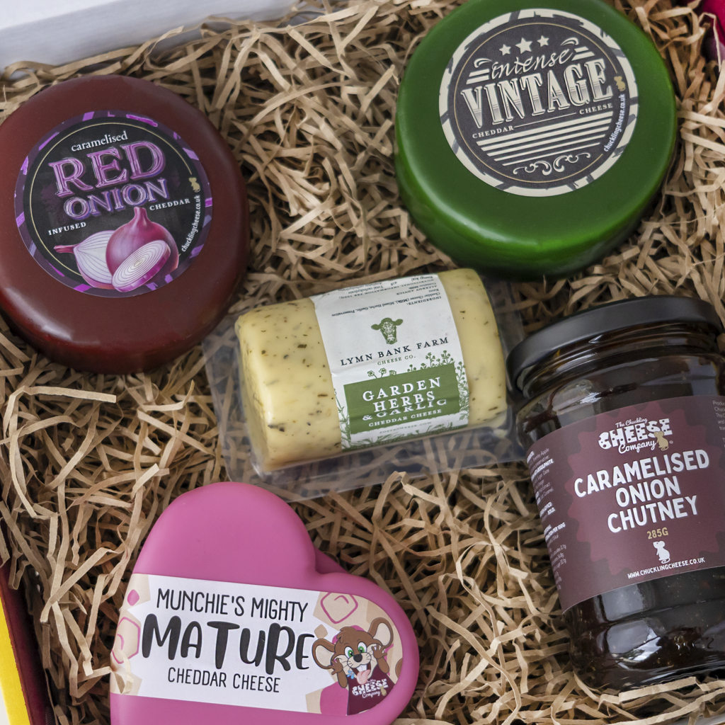 A curated cheese and chutney gift box filled with shredded paper straw, featuring multiple cheeses and chutney jars with colorful labels including red onion, vintage-style cheddar, garden herbs, caramelised onion chutney, and mature cheddar.