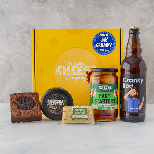 A curated gift box set from The Chuckling Cheese Company on a light marble surface against a textured grey wall. The set includes a yellow branded gift box, a chocolate-covered biscuit, a jar of cheddar spread, a stick of cheddar cheese, a jar of fart starters, and a bottle of Cranky Sod sour drink.