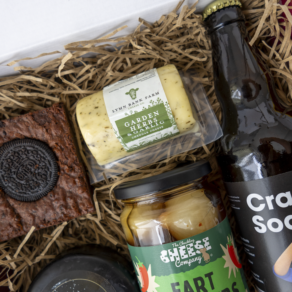 A curated snack and drink gift box filled with shredded paper. Visible items include a block of Lymn Bank Farm Garden Herb Garlic cheese, a jar of The Chuckling Cheese Company pickle onion, a brown snack bar topped with an Oreo cookie, a dark bottle of craft sod, and another dark cheese truckle.