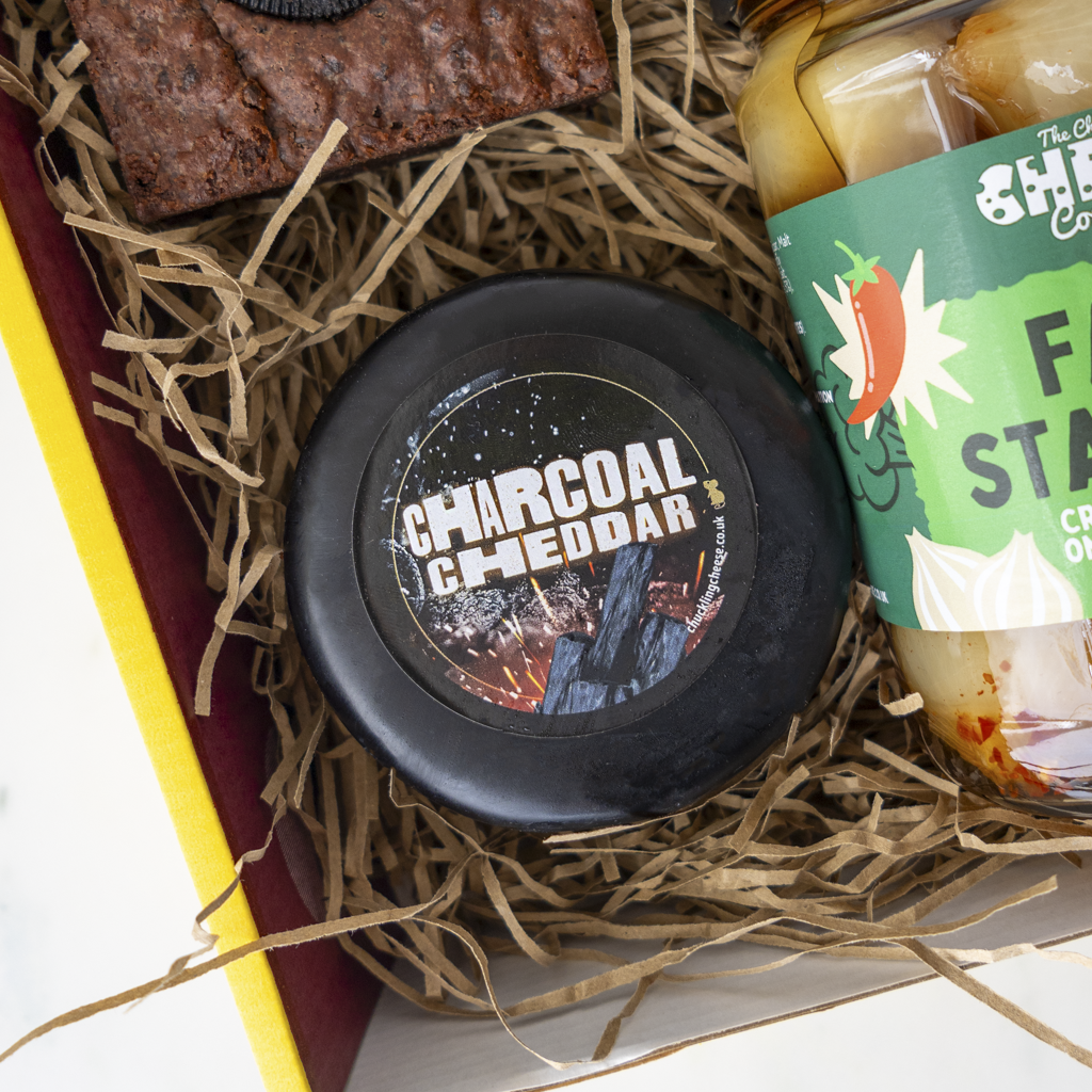 A round black cheese labeled 'CHARCOAL CHEDDAR' sits in brown paper shred inside a gift box, next to a green-labeled pickle jar and a dark brown snack piece.