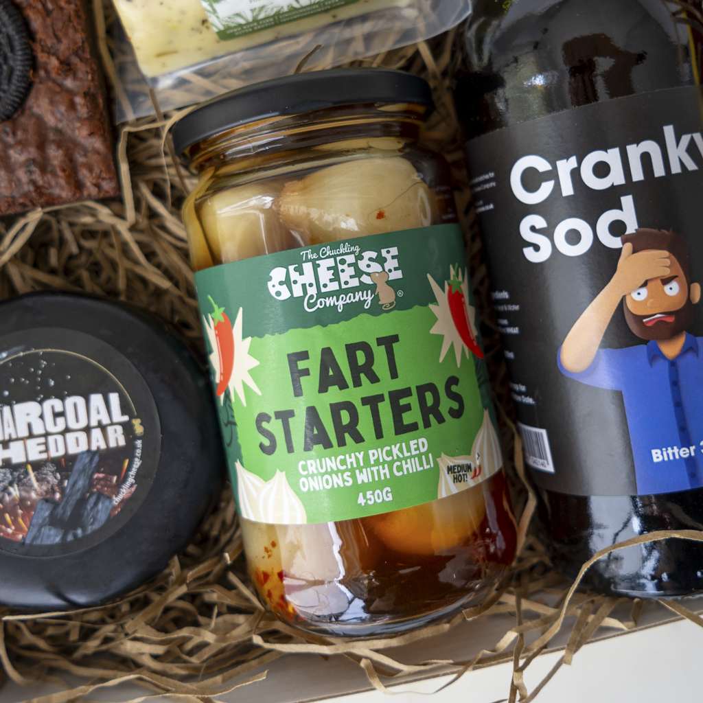 A gift box arrangement featuring a jar of 'Fart Starters' crunchy pickled onions with chilli from The Chuckling Cheese Company, a bottle of 'Crank Sod' bitter soda, and a round cheese labelled 'Charcoal Cheddar'.