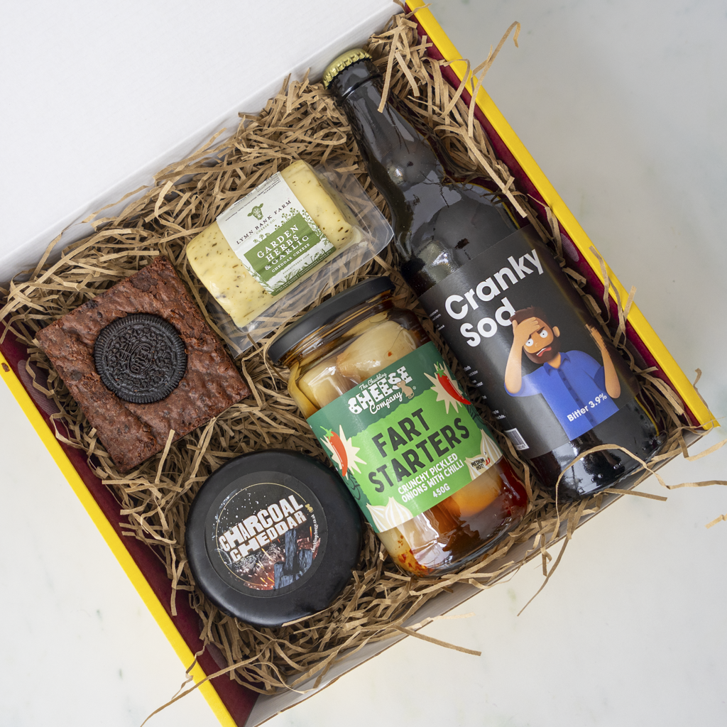 A curated gift box filled with quirky snacks and drinks, including a bottle of Cranky Sod, a jar of Fart Starters, a chocolate cookie, and a small black tin, arranged in shredded paper inside a yellow and red box.