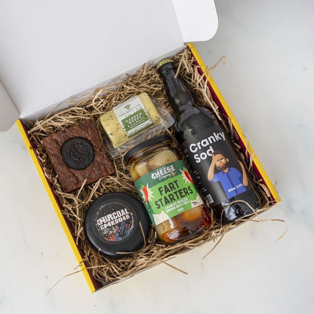 A curated gourmet gift box filled with snacks and drinks, photographed from above on a light marble surface. The box contains a chocolate bar with a round cookie on top, a small cheese wedge, a jar of pickle starters, a black truckle labelled 'Charcoal Cheddar', and a dark bottle labelled 'Cranky Sod'.