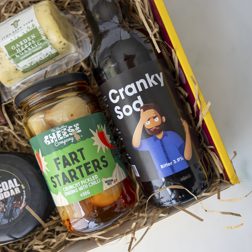 A curated gift box filled with quirky snacks and drinks, featuring a bottle of 'Cranky Sod', a jar of 'Fart Starters' pickle, a package of 'Garden Herbs & Garlic', and a dark cheese truckle in a straw-lined box.