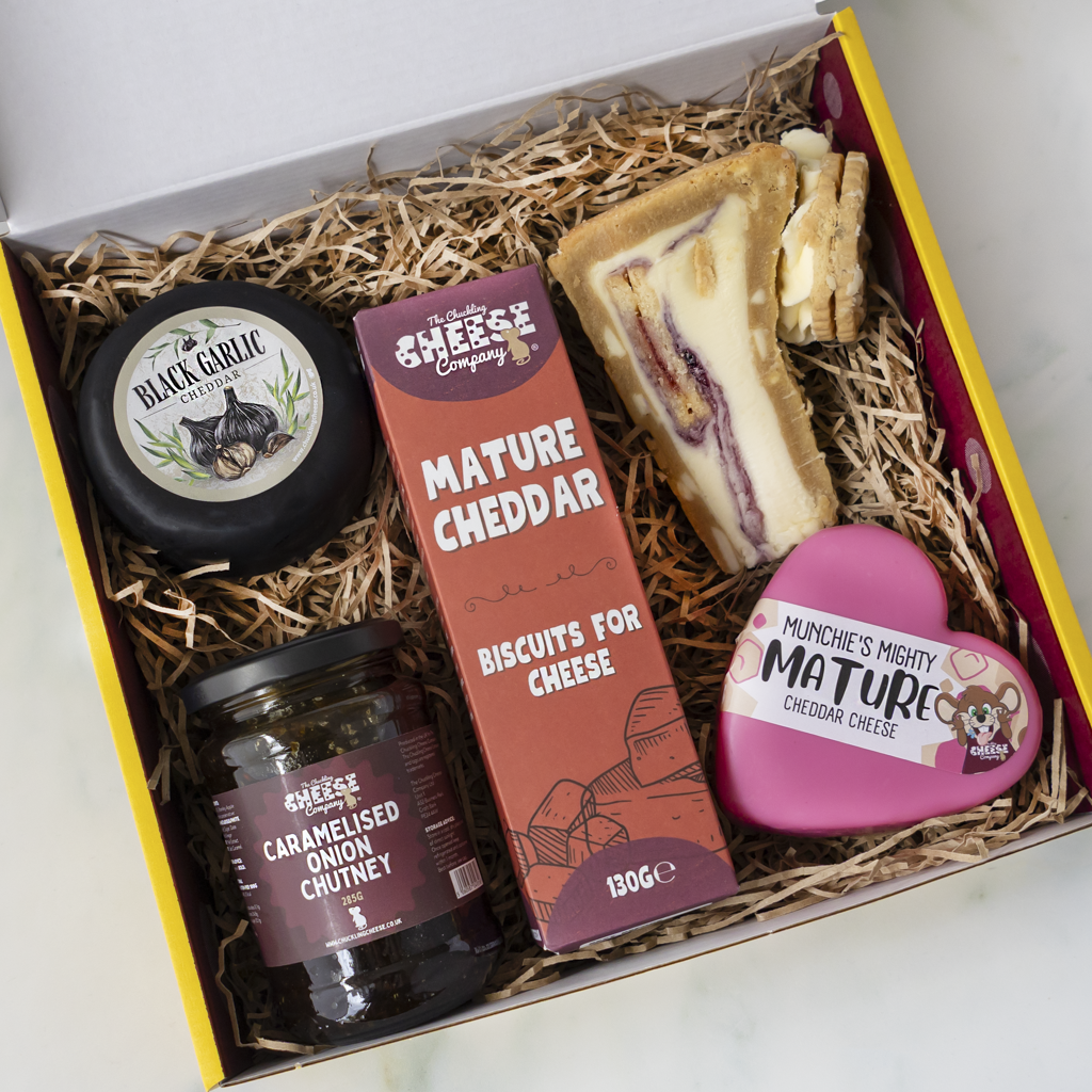 A curated cheese and cookie pie gift box filled with shredded paper. The box contains a round black cheese labeled Black Garlic, a jar of caramelised onion chutney, a box of Cheddar Biscuits, a slice of layered cookie pie, and a pink heart-shaped tin of Munchie's Mature Cheddar Cheese.