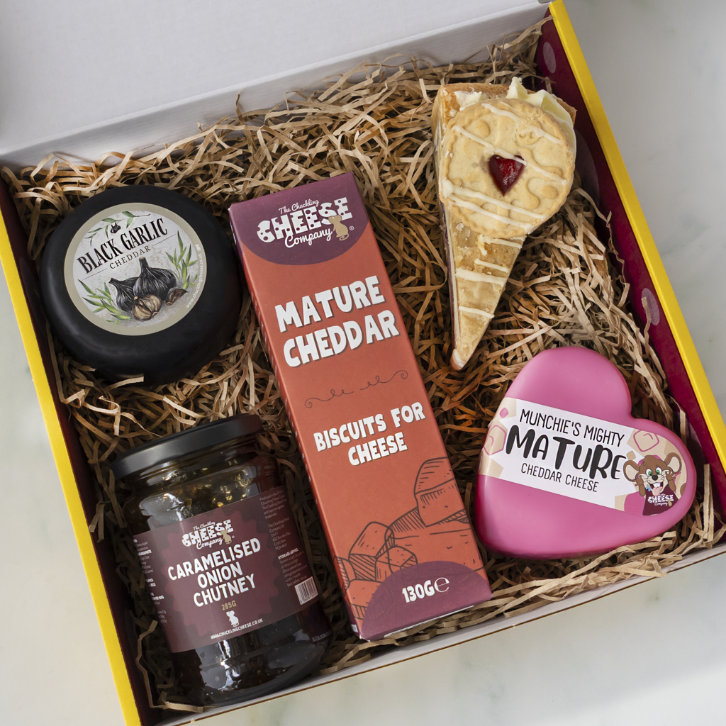 A curated cheese and snack gift box filled with shredded paper, containing a black garlic cheese wheel, a jar of caramelised onion chutney, a box of mature cheddar biscuits, a slice of cookie pie with a red heart, and a pink heart-shaped cheese truckle.