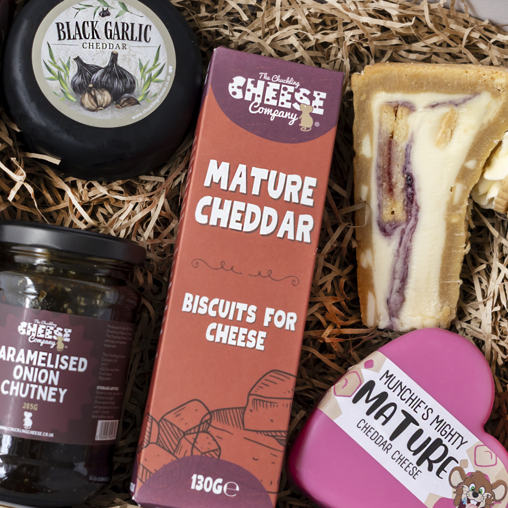 A cheese and biscuit gift basket from The Chuckling Cheese Company, arranged in a bed of shredded paper. The basket contains a round black garlic cheddar cheese wheel, a jar of caramelised onion chutney, a box of mature cheddar biscuits for cheese, a slice of cheddar sandwich, and a pink heart-shaped chocolate cheddar.