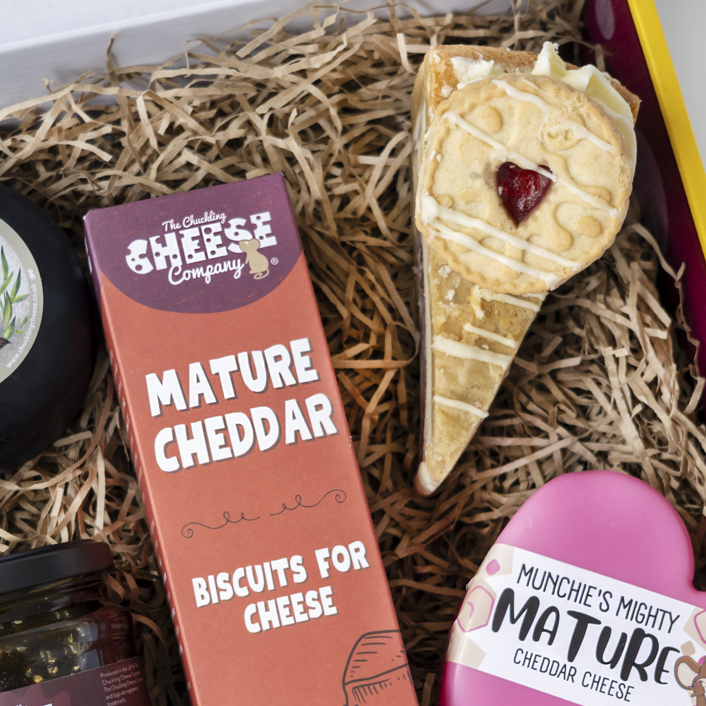 A curated cheese and biscuit gift box from The Chuckling Cheese Company, featuring a box of Mature Cheddar Biscuits for Cheese, a slice of cheddar cheese in an ice cream cone, and a pink heart-shaped cheddar cheese cookie, all nestled in brown paper shred.