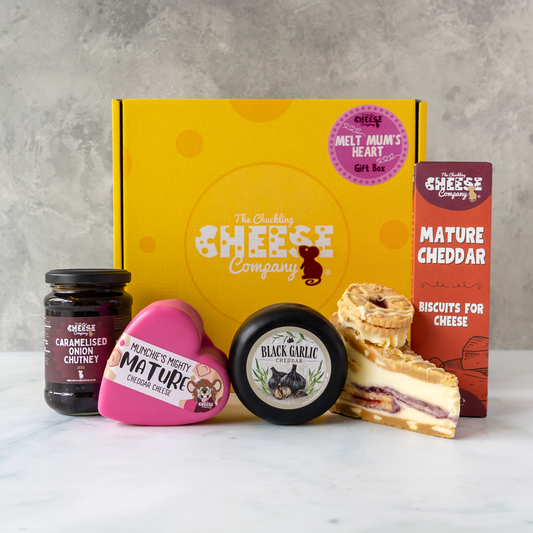 A cheese gift box set from The Chuckling Cheese Company arranged on a white marble surface against a textured grey wall. The set includes a yellow branded gift box, a jar of caramelised onion chutney, a pink heart-shaped cheese truckle, a round black garlic cheese truckle, and a slice of layered cookie pie and cheese biscuits.