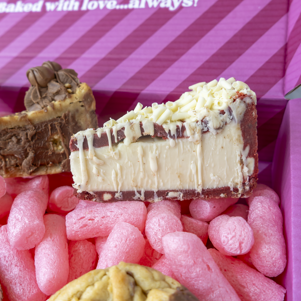 A close-up of a slice of chocolate and cream cookie pie with white chocolate drizzle inside a pink gift box with other baked goods partially visible.