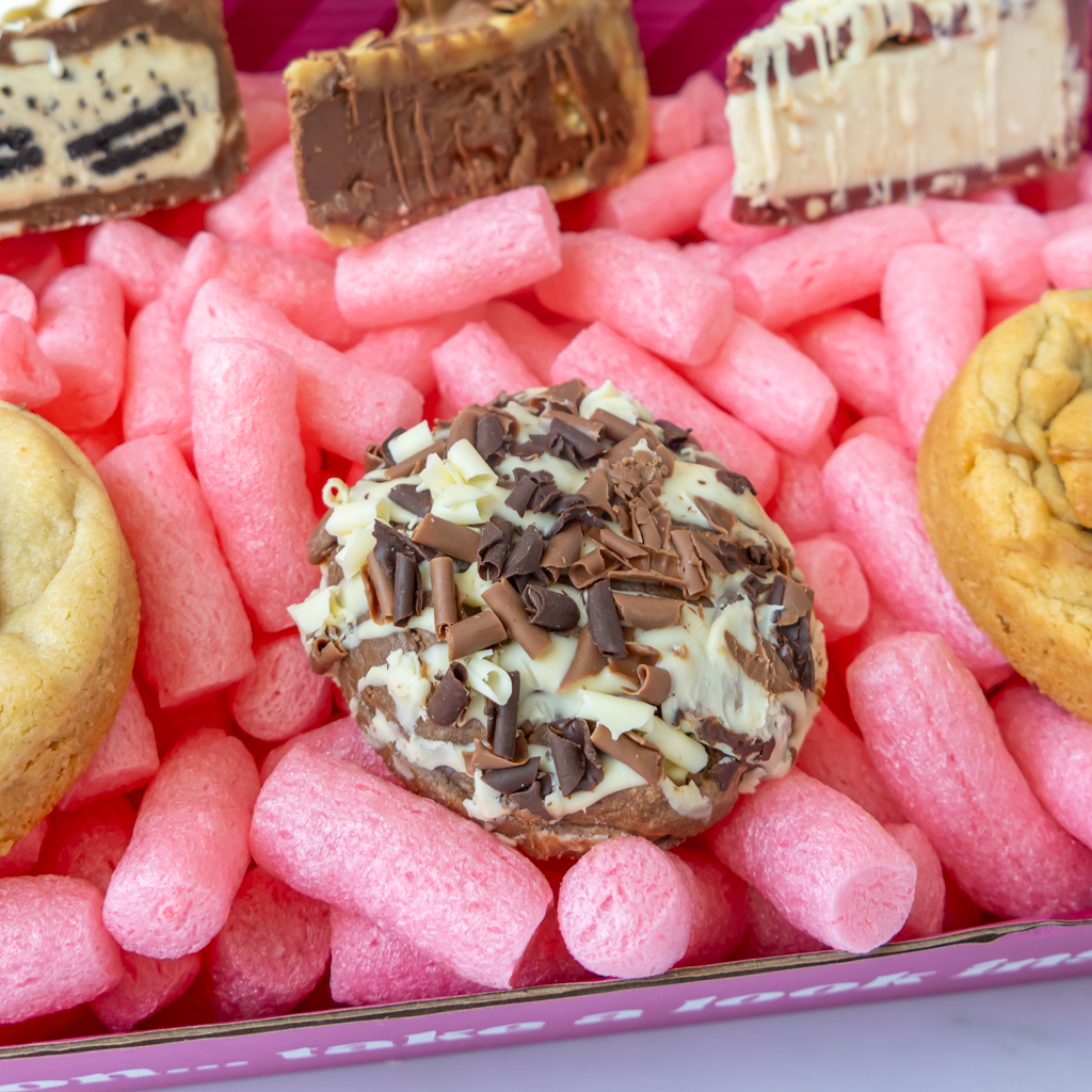A close-up of a boxed snack assortment filled with pink bubble wrap. In the center is a round cookie topped with white chocolate and chocolate shavings, surrounded by other cookies.