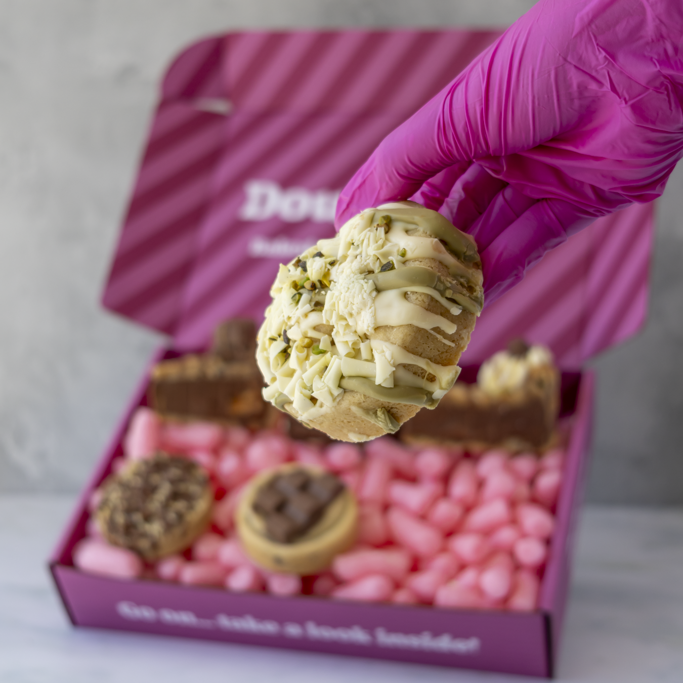 A hand wearing a bright pink glove holds a decorated cookie above an open pink gift box filled with chocolate treats. The box lid has partial white branding text "DoughGirl".
