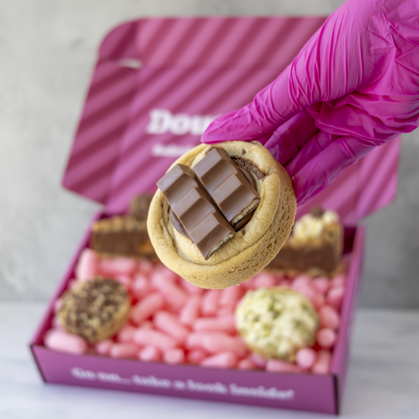 A hand wearing a bright pink glove holds a chocolate-covered cookie in front of an open pink gift box filled with assorted chocolates. The box lid has partial white branding text that appears to read 'DoughGirl".