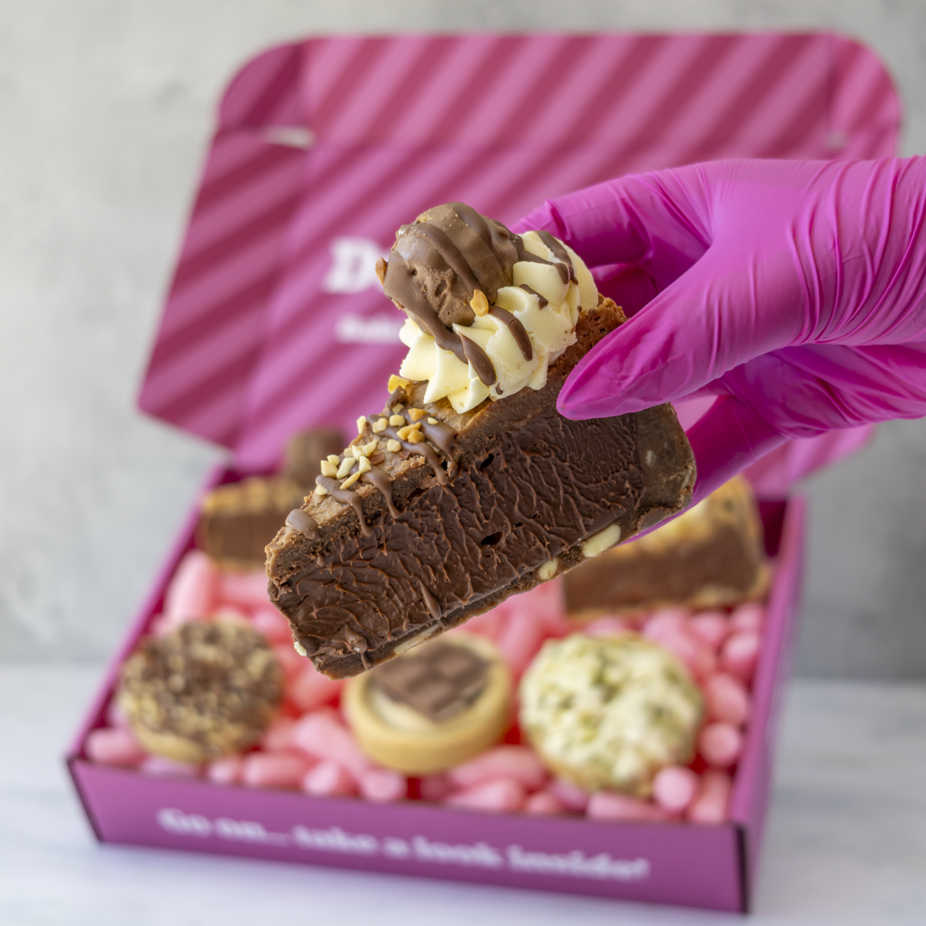 A hand wearing a bright pink glove holds a chocolate-covered dessert slice in front of an open pink gift box filled with assorted treats.