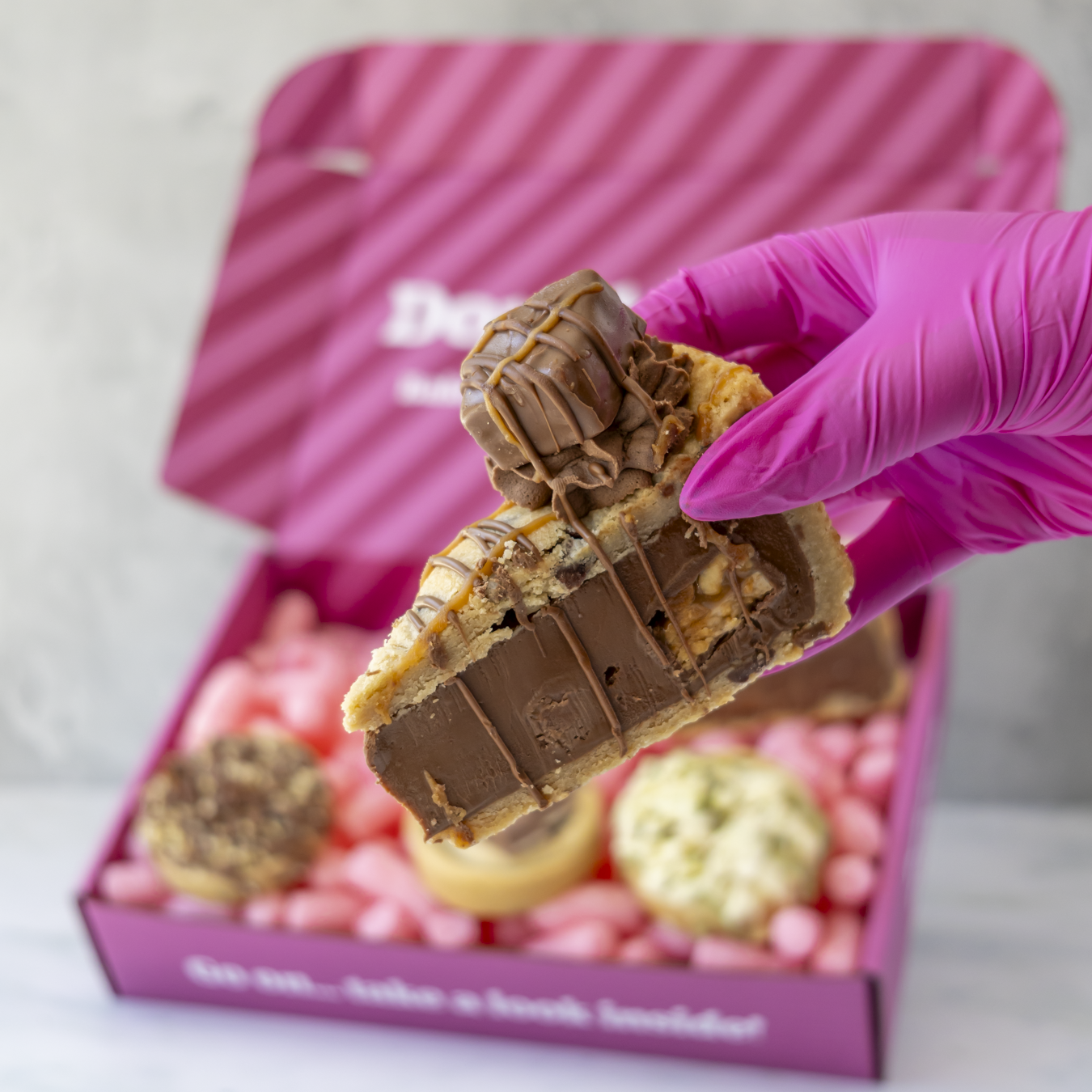 A hand wearing a bright pink glove holds a chocolate-dipped cookie pie slice in front of an open pink gift box filled with assorted cookies.