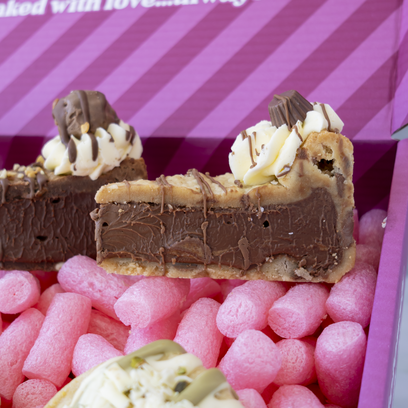 Chocolate and cream pie slices displayed in a pink gift box filled with pink packing peanuts. The box has a purple and pink striped lid with partial text reading.