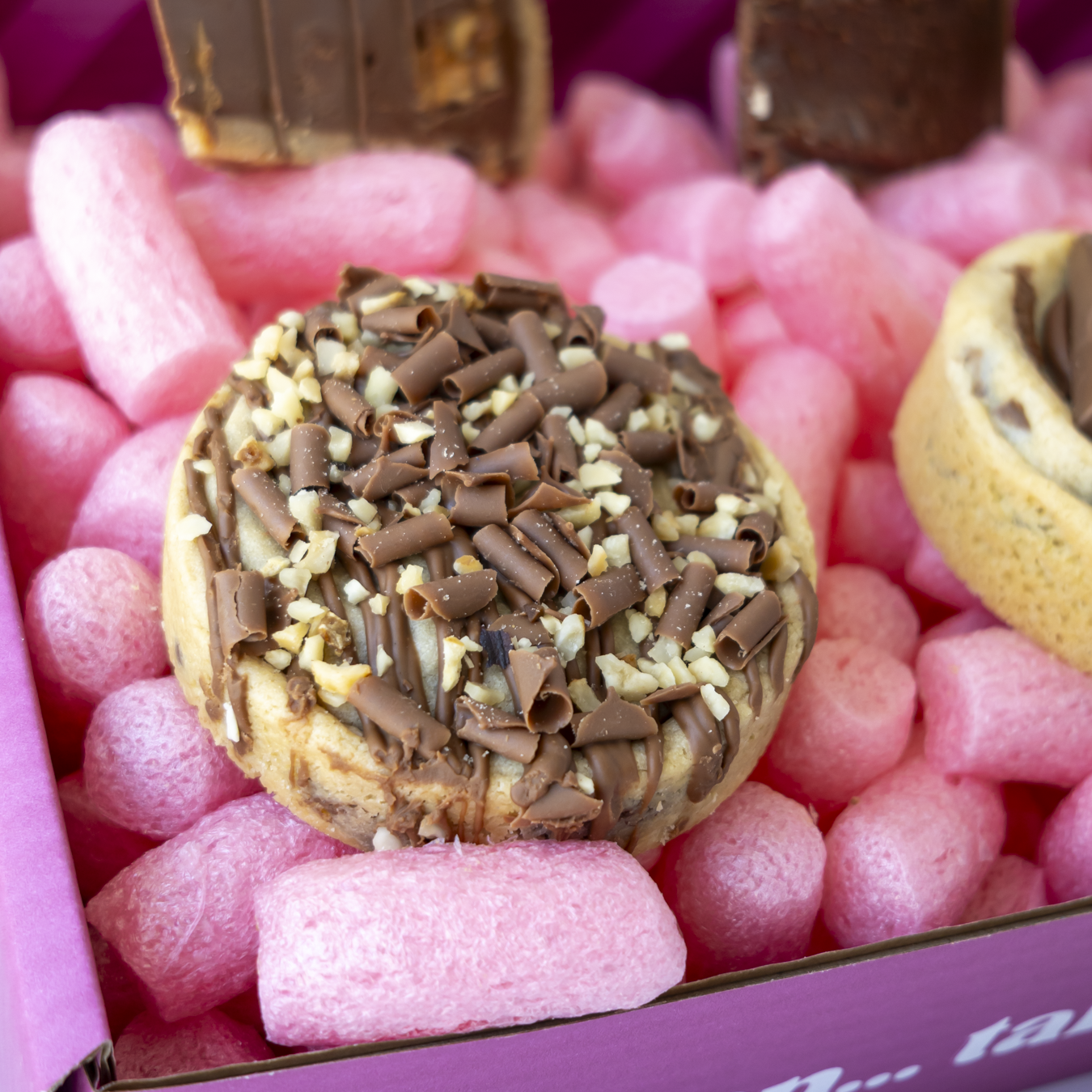 A close-up of a chocolate-covered cookie or pastry topped with chocolate shavings and crushed nuts,inside a pink box.