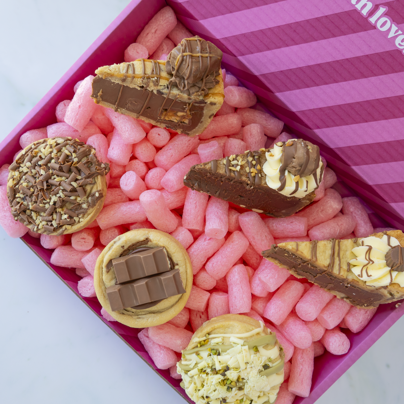 A gift box of assorted gourmet chocolate treats, including chocolate cake slices, chocolate-covered cookies, and a chocolate bar, arranged on pink packing peanuts inside a pink striped box.